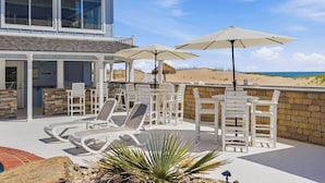 Ein zweistöckiges Haus mit einer Terrassenanlage, ausgestattet mit weißen Liegestühlen, Sonnenschirmen und einem Pool.