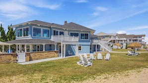 Ein zweistöckiges Strandhaus mit überdachter Terrasse, Gartenmöbeln und einer Treppe zu einem Balkon.