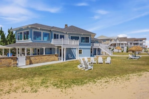 A two-story beachfront house with a covered patio, outdoor seating, and a staircase leading to a balcony.