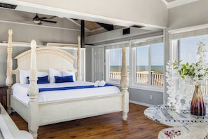 A four-poster bed with a white headboard and blue bedding, large windows with a view of the ocean, and a ceiling fan.