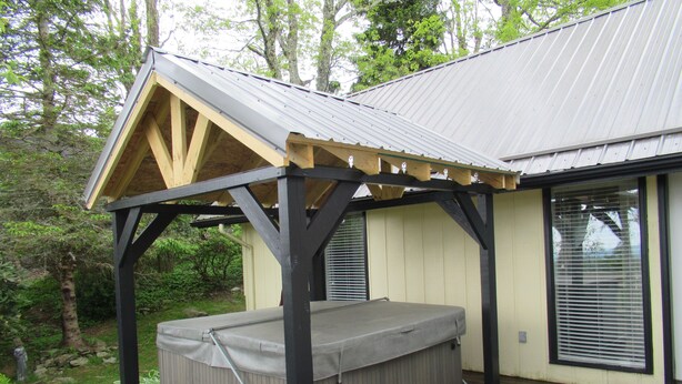 Un espace abrité avec un jacuzzi, accessible par des marches, une terrasse en bois et une maison dotée d’un toit en métal et de grandes fenêtres.