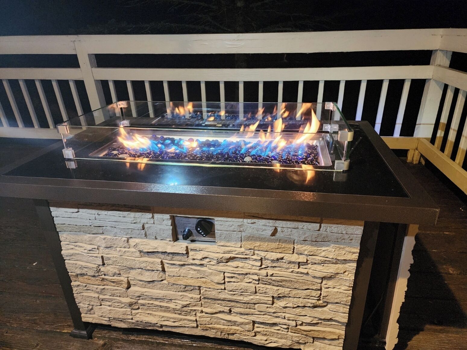 A rectangular outdoor fire feature with a glass top and stone base, surrounded by a white railing.