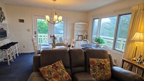 Un salon avec un canapé en cuir marron, deux coussins à motifs floraux, un lustre et un coin repas comprenant des chaises blanches et une table.