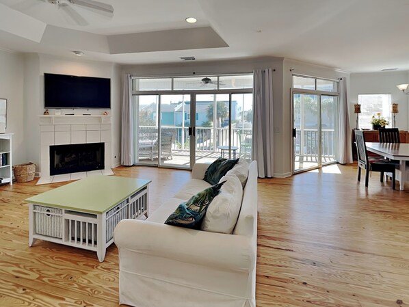 A spacious living room with a white sofa, a flat-screen TV mounted above a fireplace, and a sliding glass door leading to a balcony.