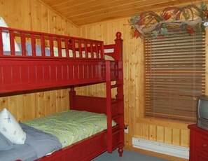 A wooden room with a bunk bed, a single bed, a television, and a window with blinds.