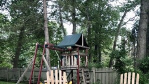 A wooden swing set, two Adirondack chairs, and a small wooden playhouse in a wooded area.