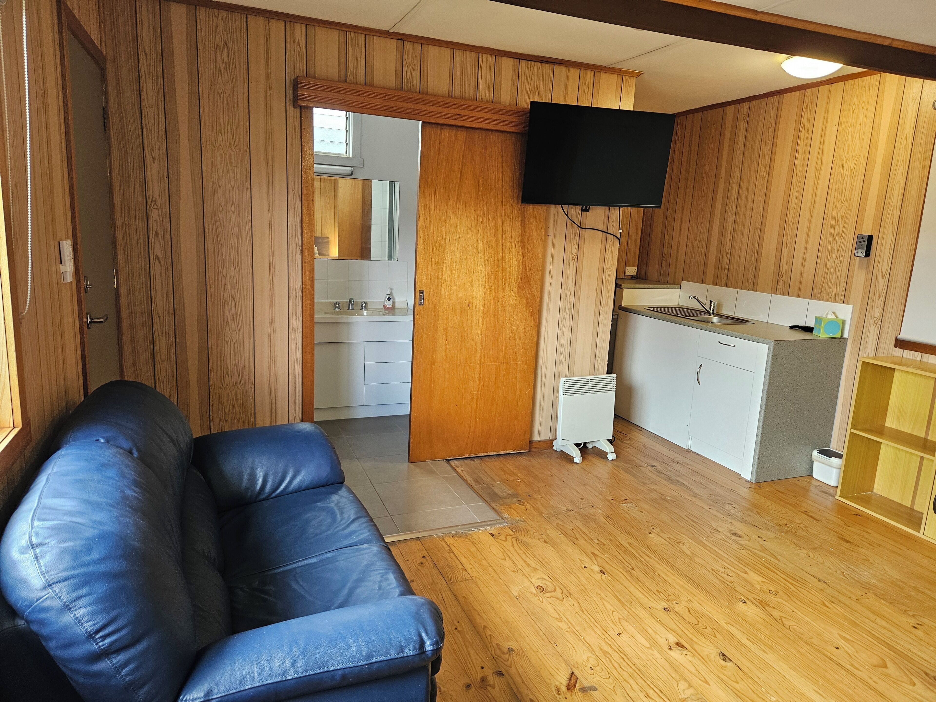 Une pièce avec un plancher en bois, un canapé en cuir bleu, une cuisine avec des meubles blancs et un téléviseur fixé au mur.