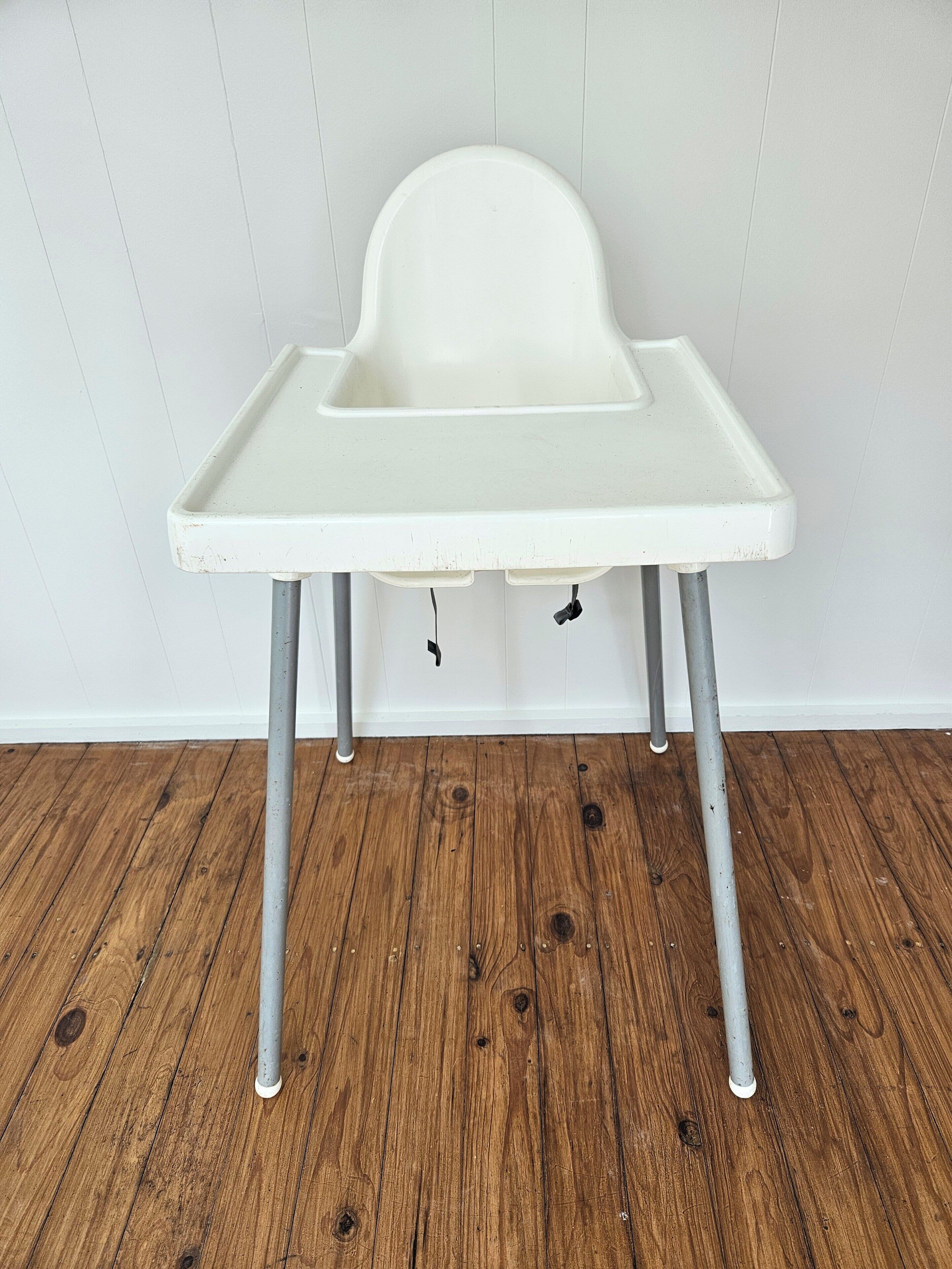 Une chaise haute blanche, dotée d’un plateau, soutenue par quatre pieds en métal, est placée contre un mur blanc, au-dessus d’un plancher en bois.