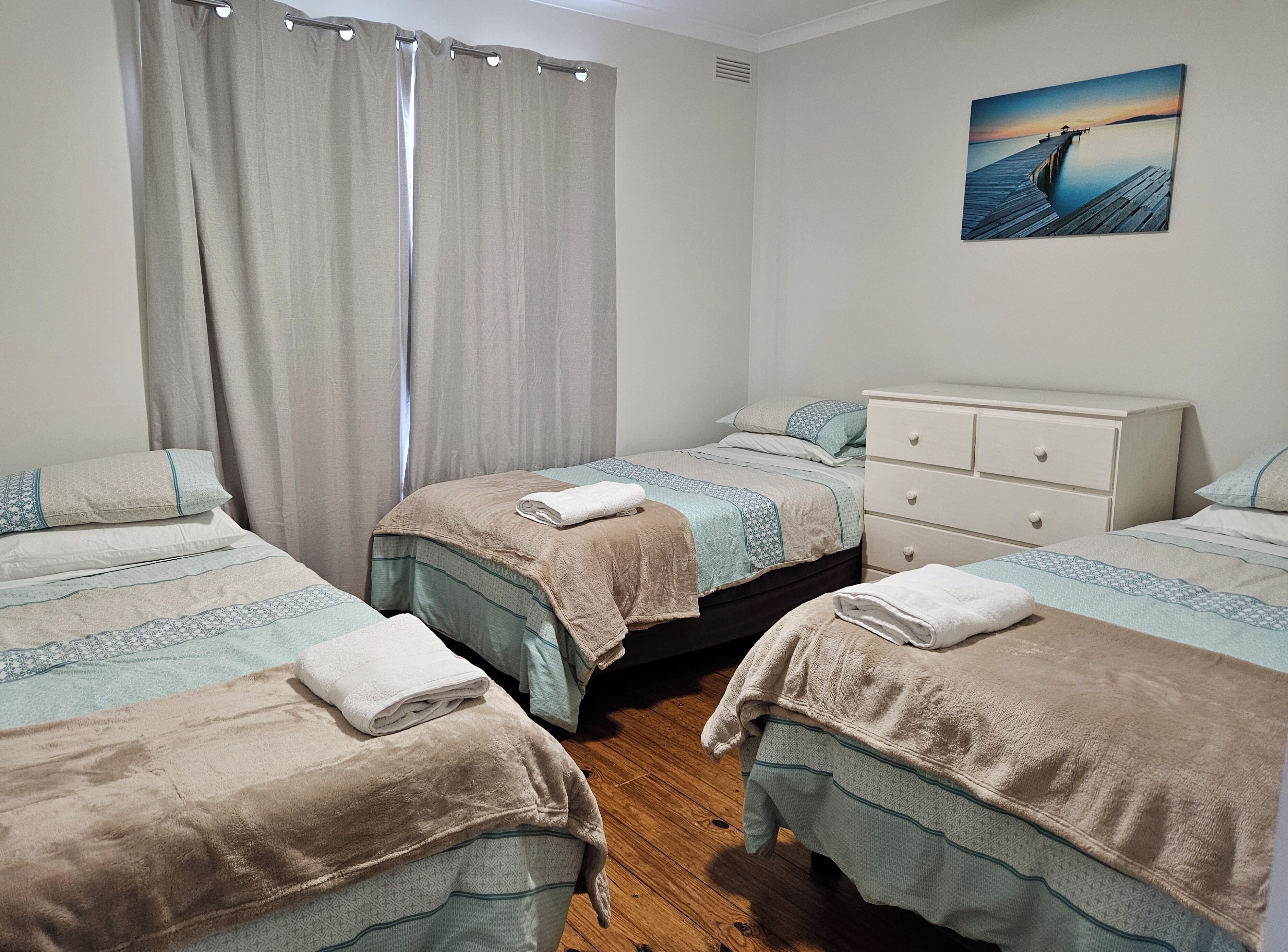 Een slaapkamer met twee bedden, een witte dressoir, een schilderij van een pier en een raam met gordijnen.