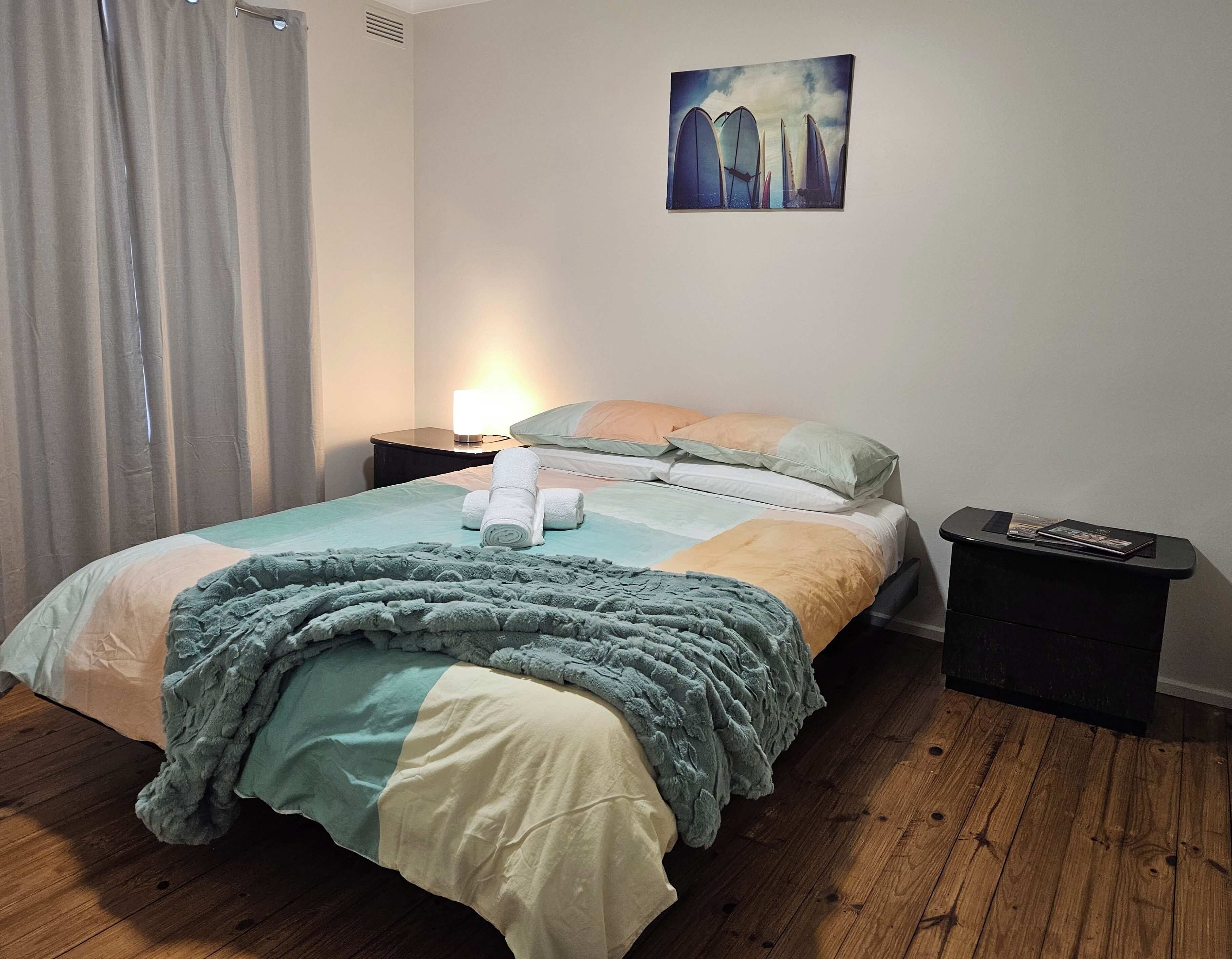 A neatly made bed with a blue blanket, a pillow, and a rolled towel. There is a nightstand with a lamp and a magazine. The room has wooden flooring and a painting on the wall.
