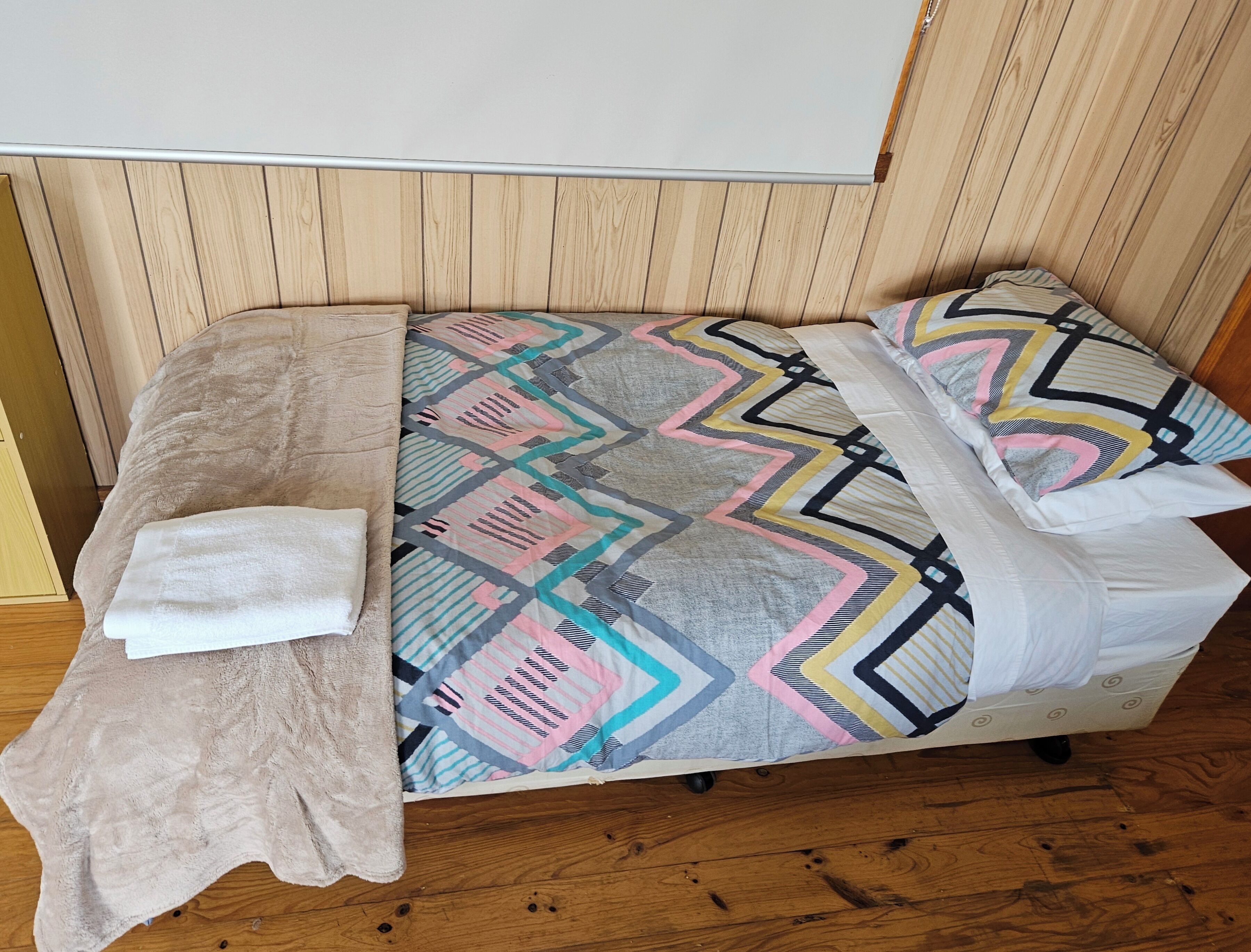 A single bed with a patterned bedspread, a white pillow, and a beige towel on the floor.