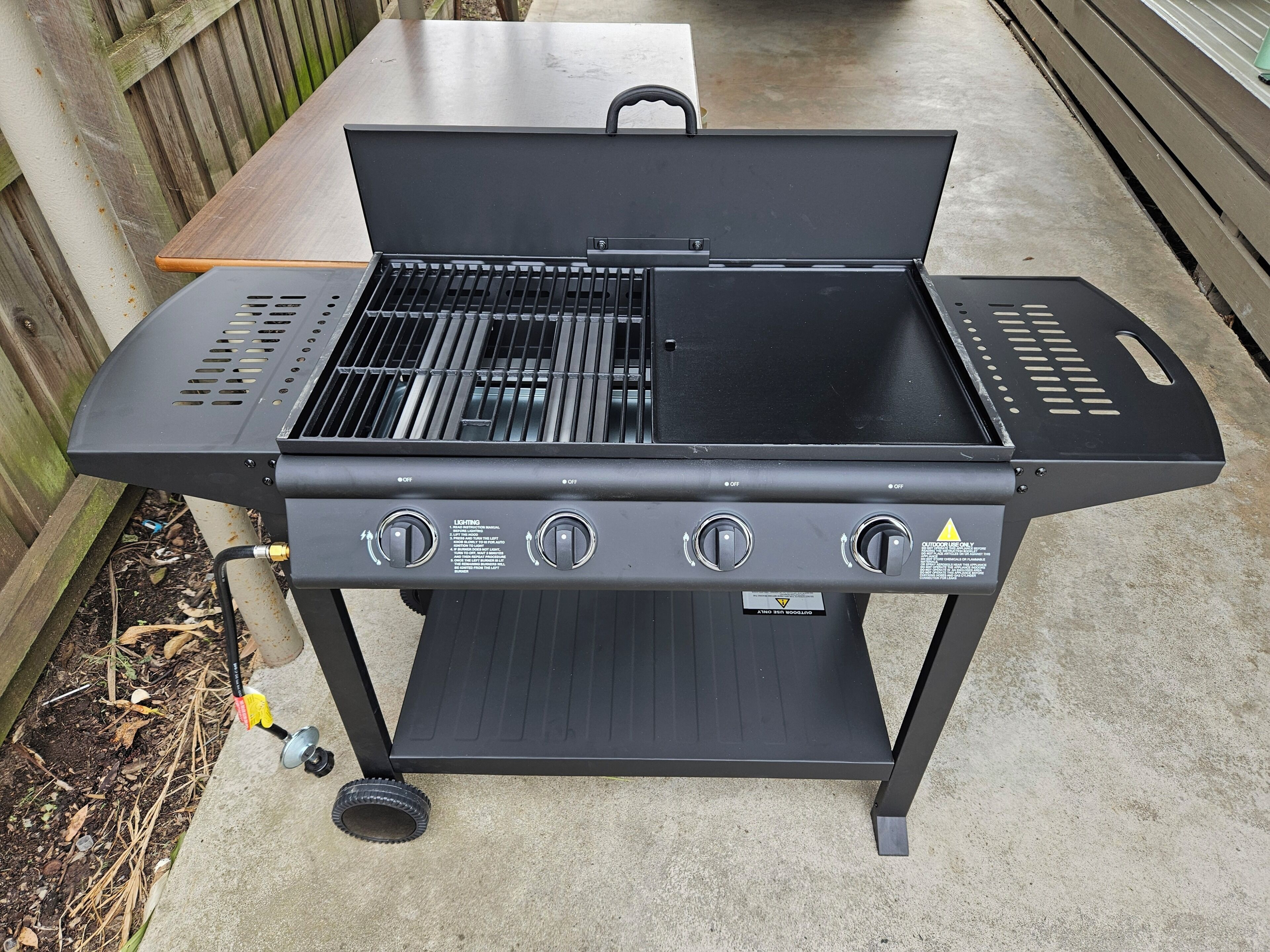 A black outdoor grill with four burners, a handle, and a gas hose.