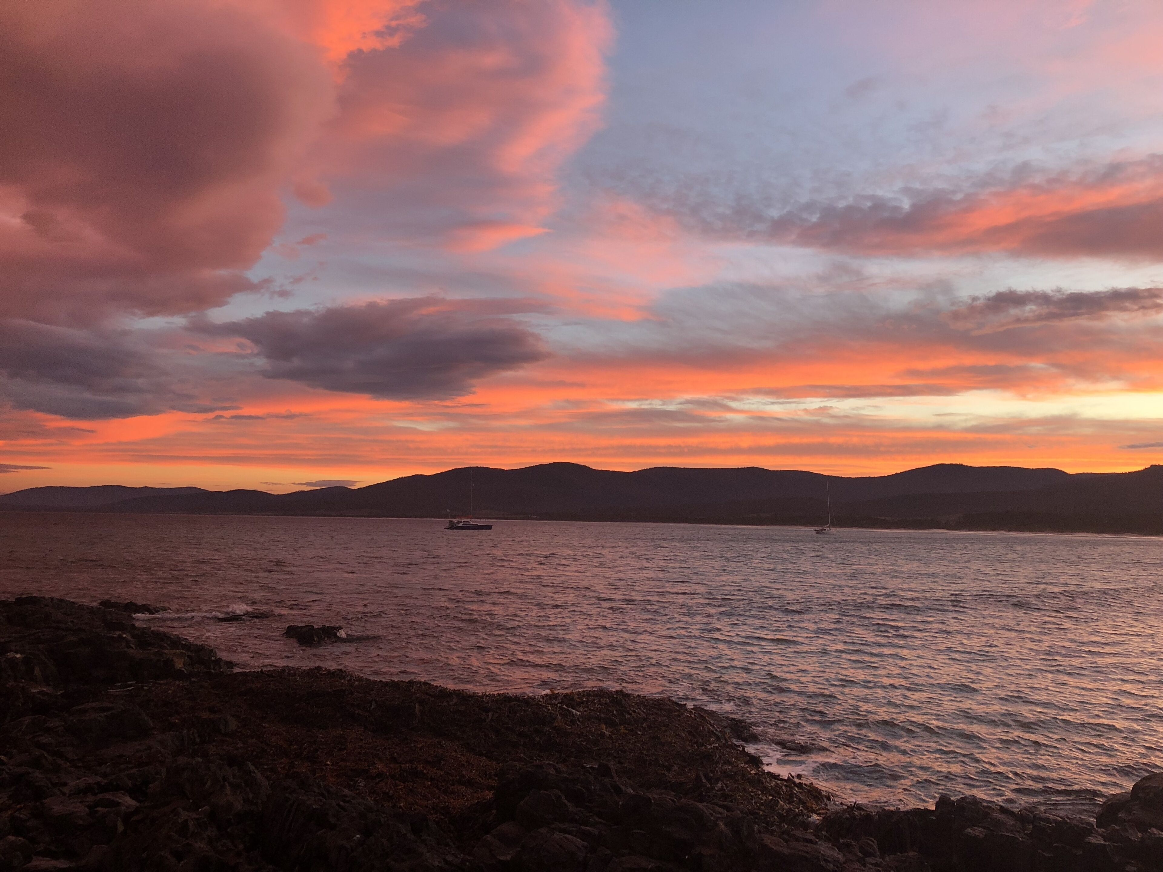 A coastal landscape with a sunset, rocky shoreline, and distant mountains.