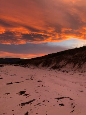 Hiekkaranta, jossa on vähän kasvillisuutta, auringonlaskun värjäämän taivaan alla.
