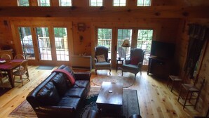 A cozy cabin interior with a leather sofa, a coffee table, and a TV. Large windows offer a view of the outdoors.
