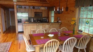 A wooden dining room with a large table, white chairs, and a kitchen in the background.