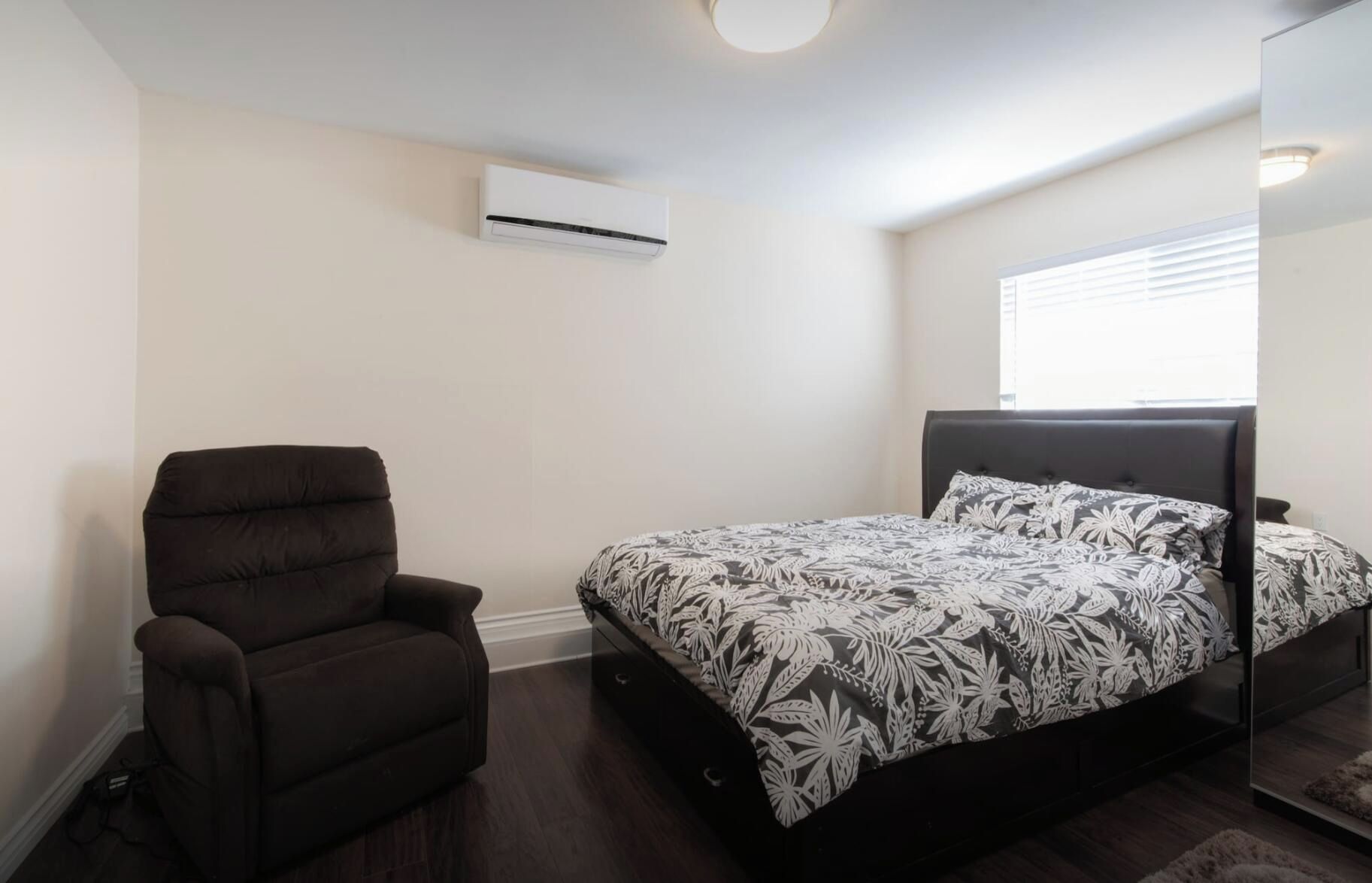 A bedroom with a bed, a chair, an air conditioning unit, and a window.