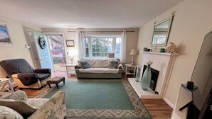 A cozy living room with a sofa, armchair, and ottoman, a fireplace, and a view of the outdoors through a large window. - Quiet 3BR Ranch - Little Dog Friendly! (2033) (Harwich)