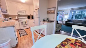 A compact kitchen with white cabinets, a microwave, and a refrigerator. There is a dining table with a colorful table runner. The room has wooden flooring and a view of the outside through a window. - Quiet 3BR Ranch - Little Dog Friendly! (2033) (Harwich)