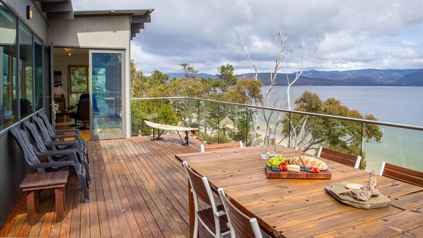 Eine Holzterrasse mit gedecktem Tisch, mit Blick auf ein Gewässer und Hügel.
