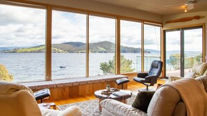 Ein geräumiges Wohnzimmer mit großen Fenstern und Blick auf einen See, ausgestattet mit Ledersofas und einem schwarzen Sessel.