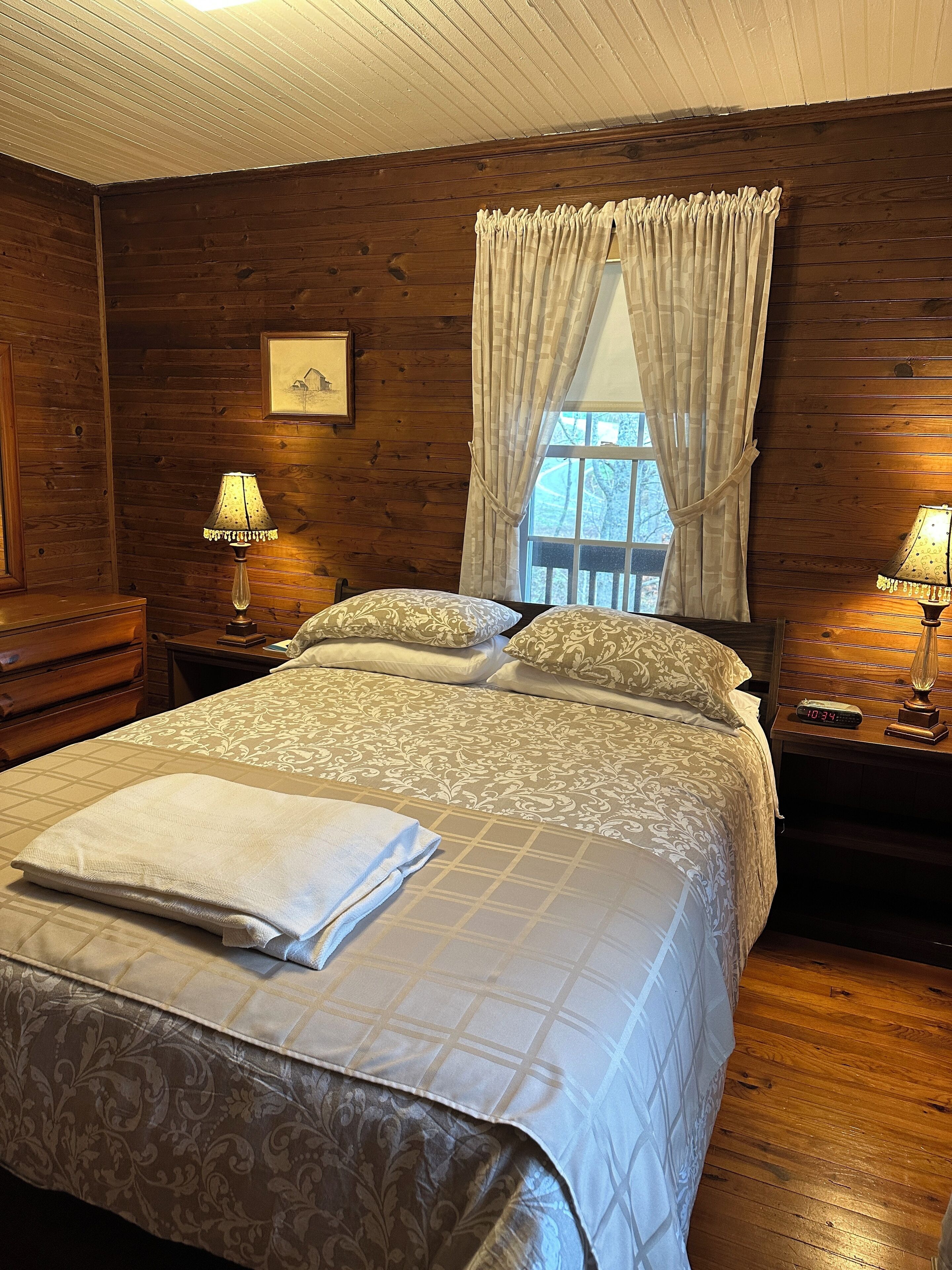 A cozy bedroom with a large bed, wooden walls, a window with curtains, and two bedside lamps.