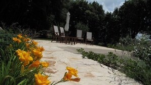 Un chemin de pierres mène à un espace extérieur aménagé avec des parasols blancs, le tout entouré de verdure et de fleurs jaunes.