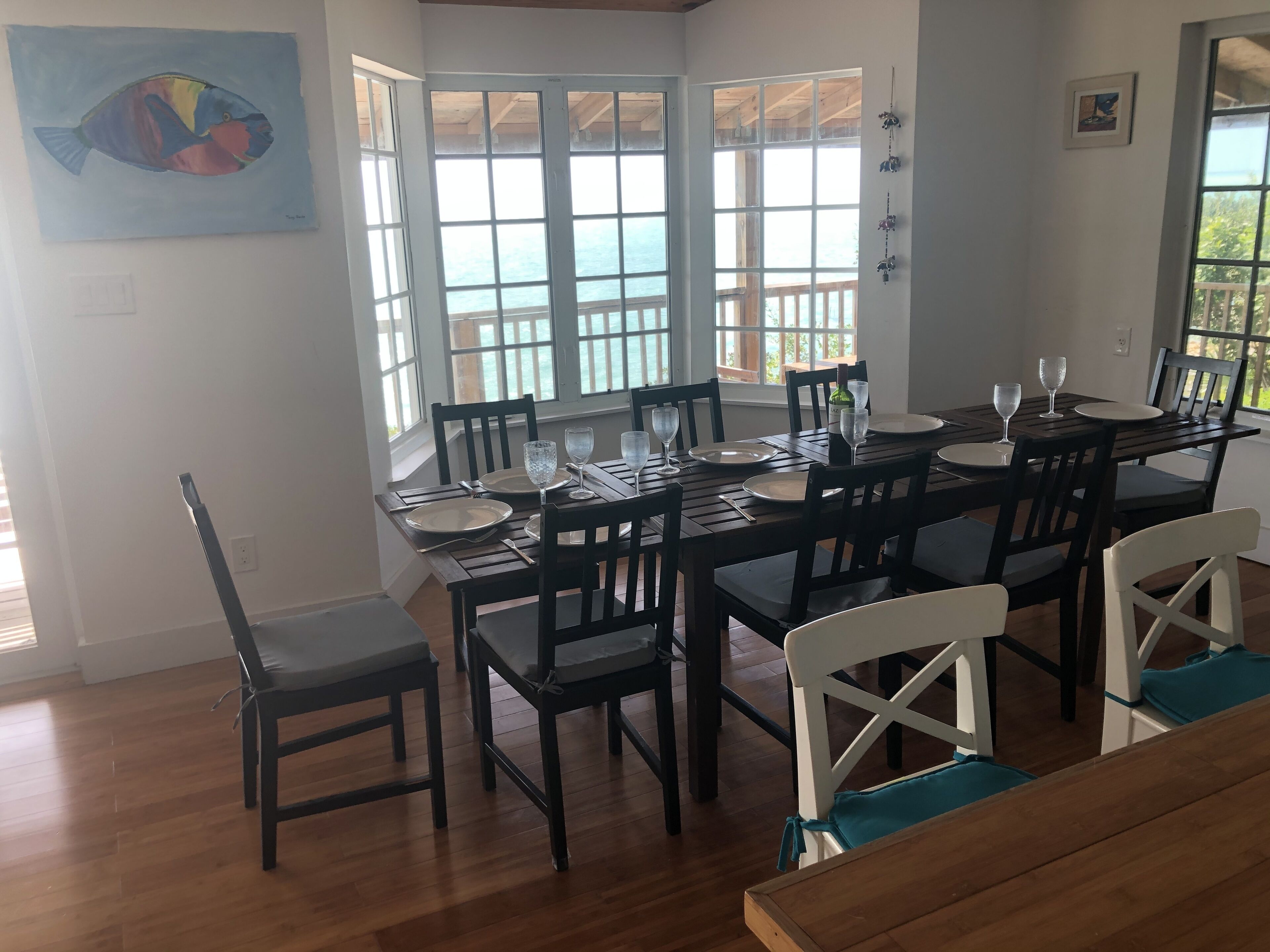 A dining area with a large table set for six, surrounded by wooden chairs, large windows with a view of the sea, and a painting on the wall.