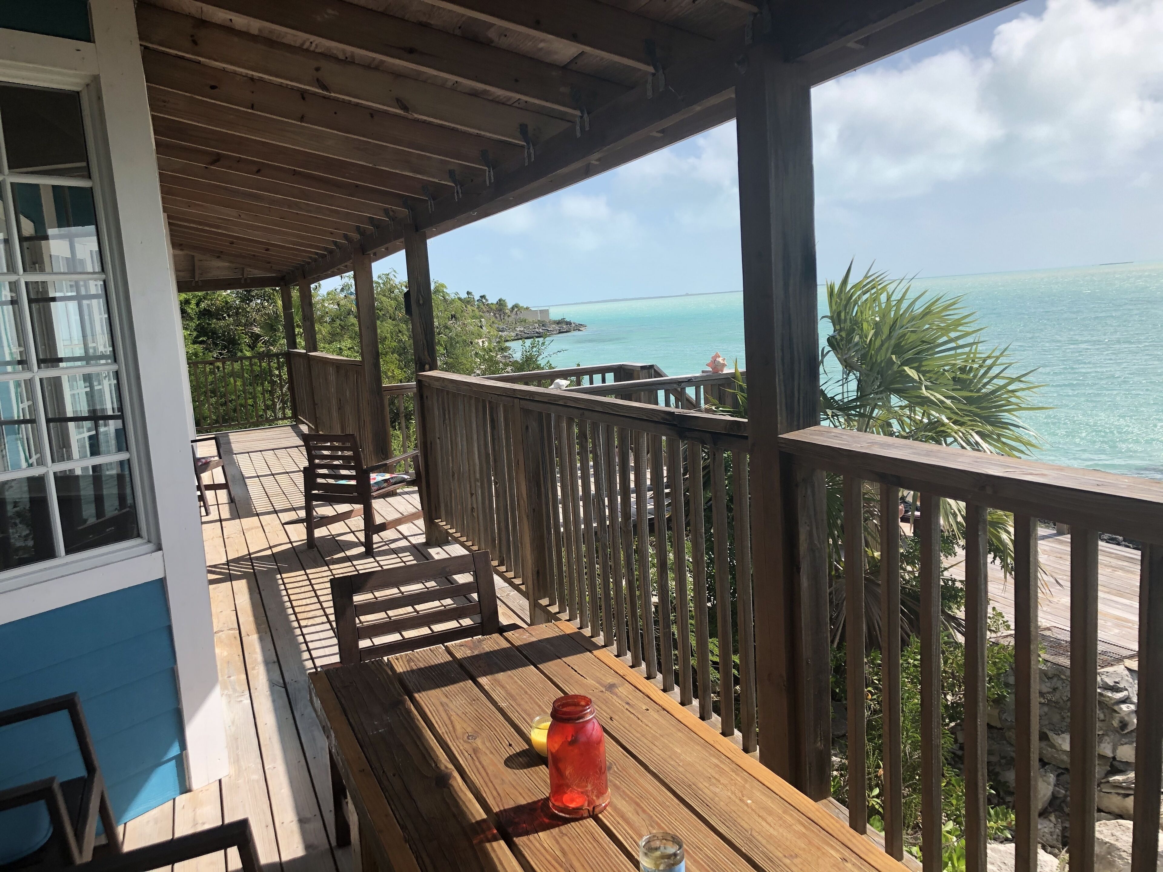 A wooden deck with a table and chairs, overlooking a clear blue sea and a rocky shoreline.