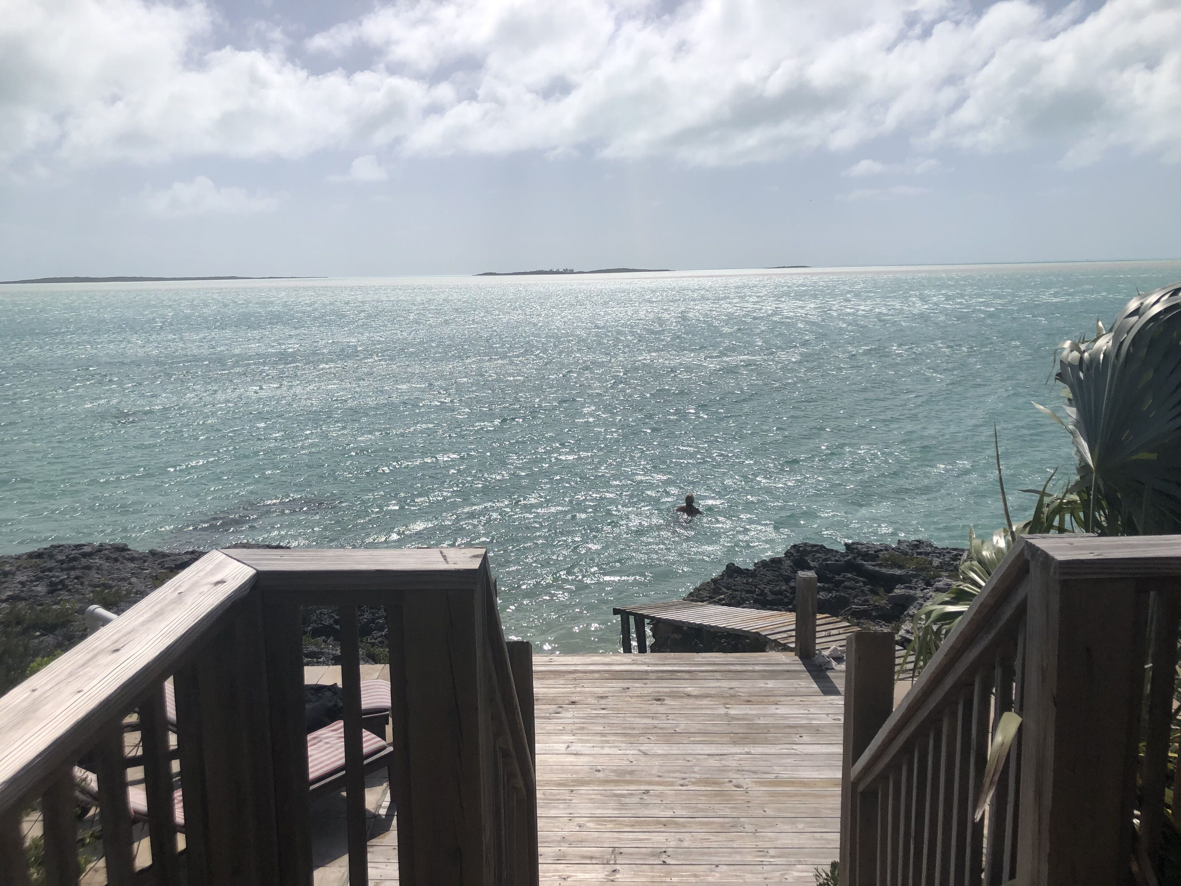 A wooden deck leading to a beach with clear water and a distant shoreline.