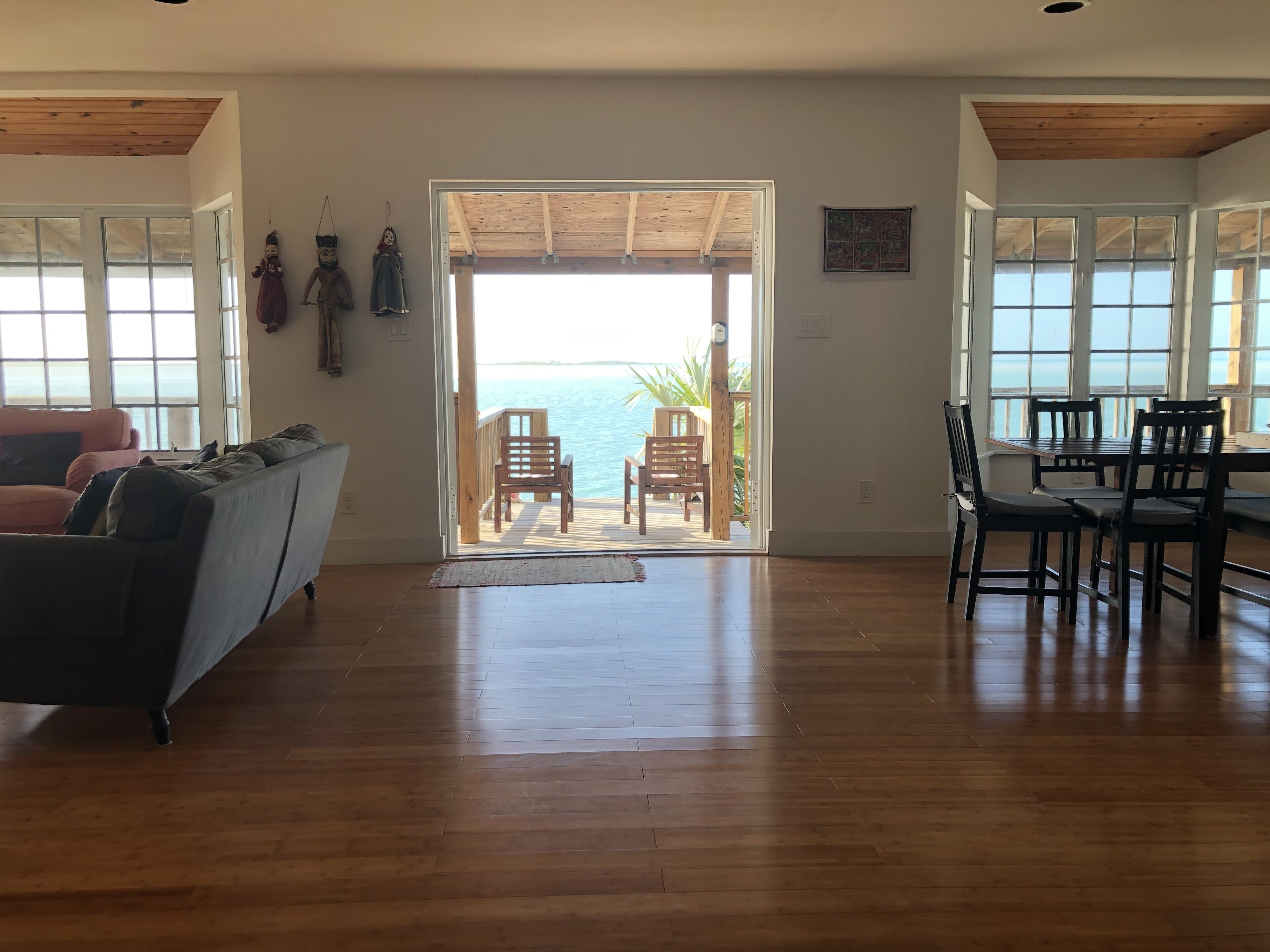 A spacious living area with a wooden floor, a sofa, a dining table with chairs, and a view of the sea through a large glass door.