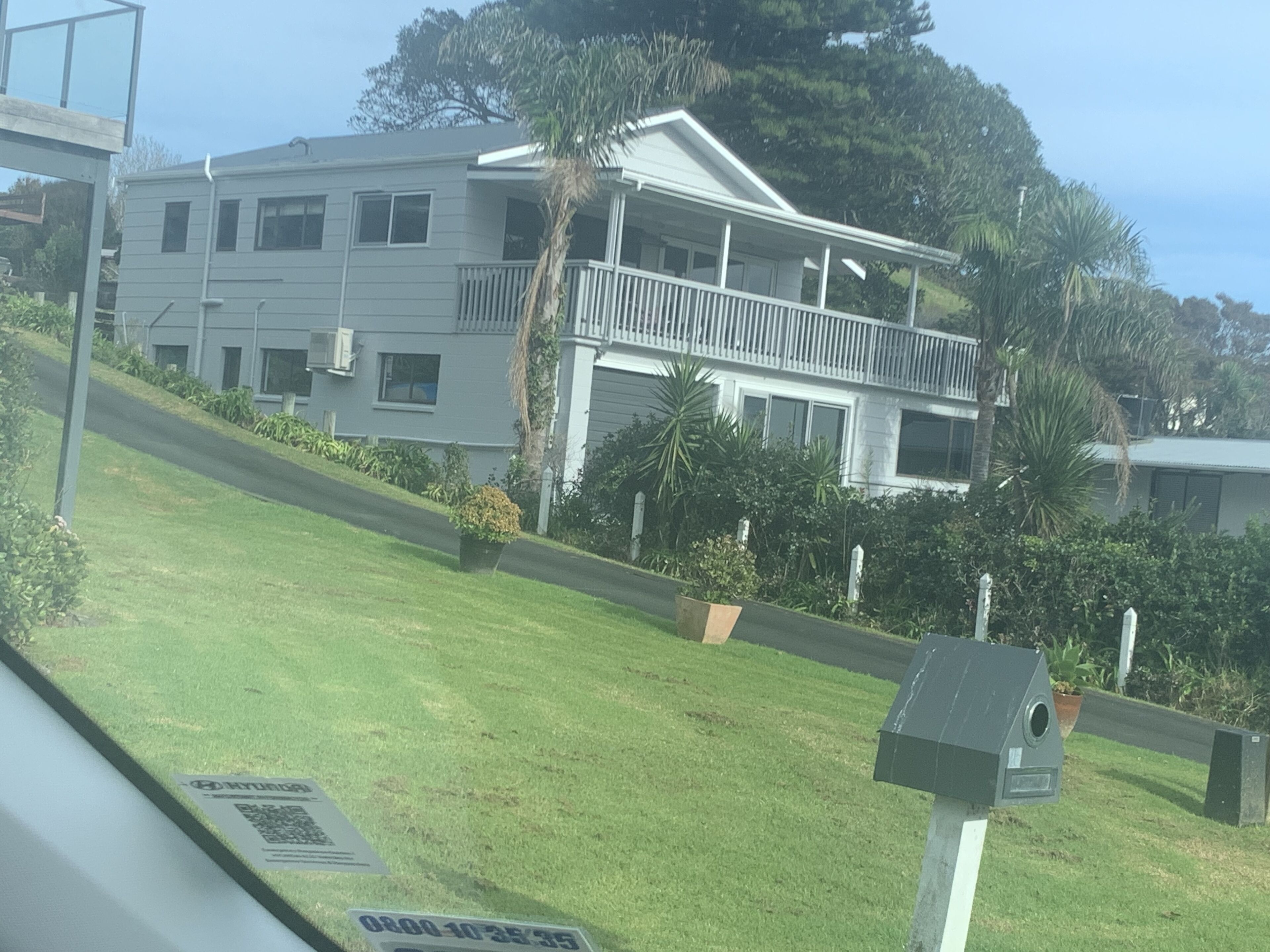 Un edificio de dos pisos con un balcón, rodeado de vegetación y un cielo despejado.
