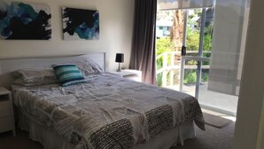 A bedroom with a large bed, a nightstand, a lamp, and a view of the outdoors through a sliding door. - Teaghlach: Tapeka / Russell Holiday Home (Tapeka Point (Russell))