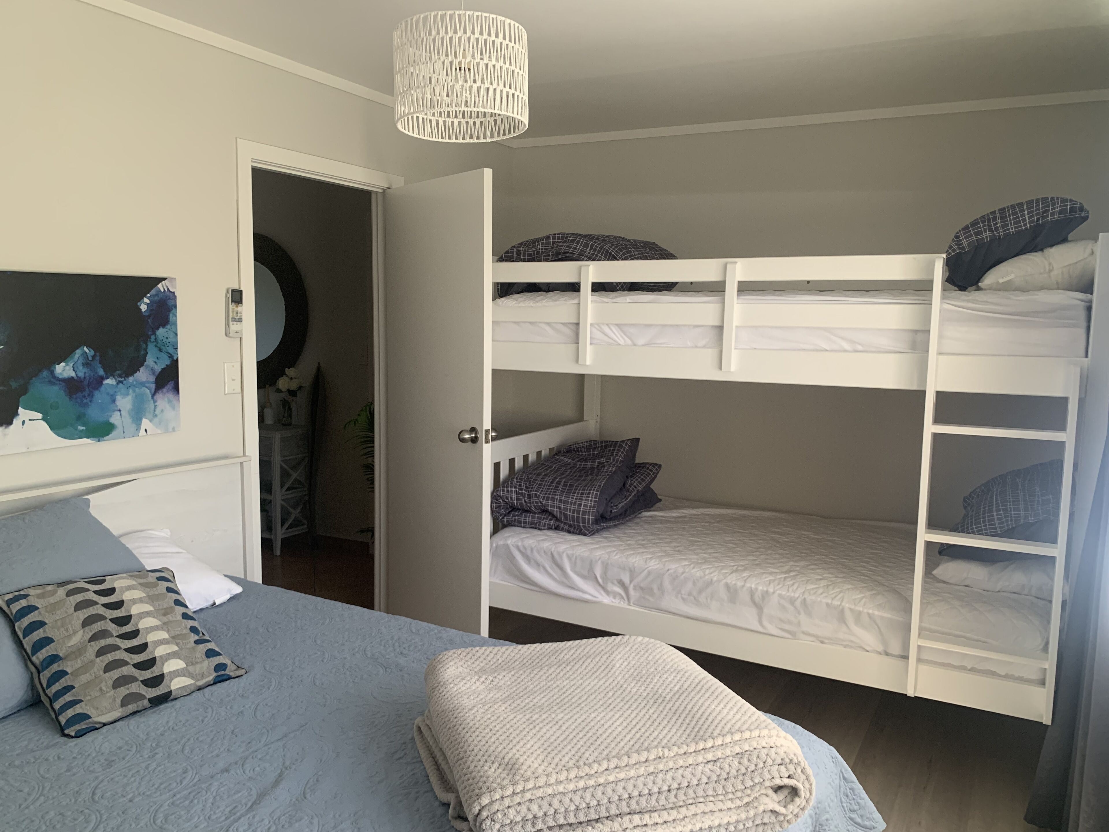 A room with a bunk bed, a bedside table, a chair, a painting, and a door leading to another room.