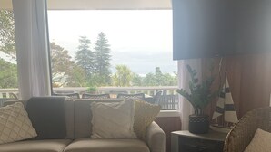 A living room with a sofa, a coffee table, and a view of greenery outside. - Teaghlach: Tapeka / Russell Holiday Home (Tapeka Point (Russell))