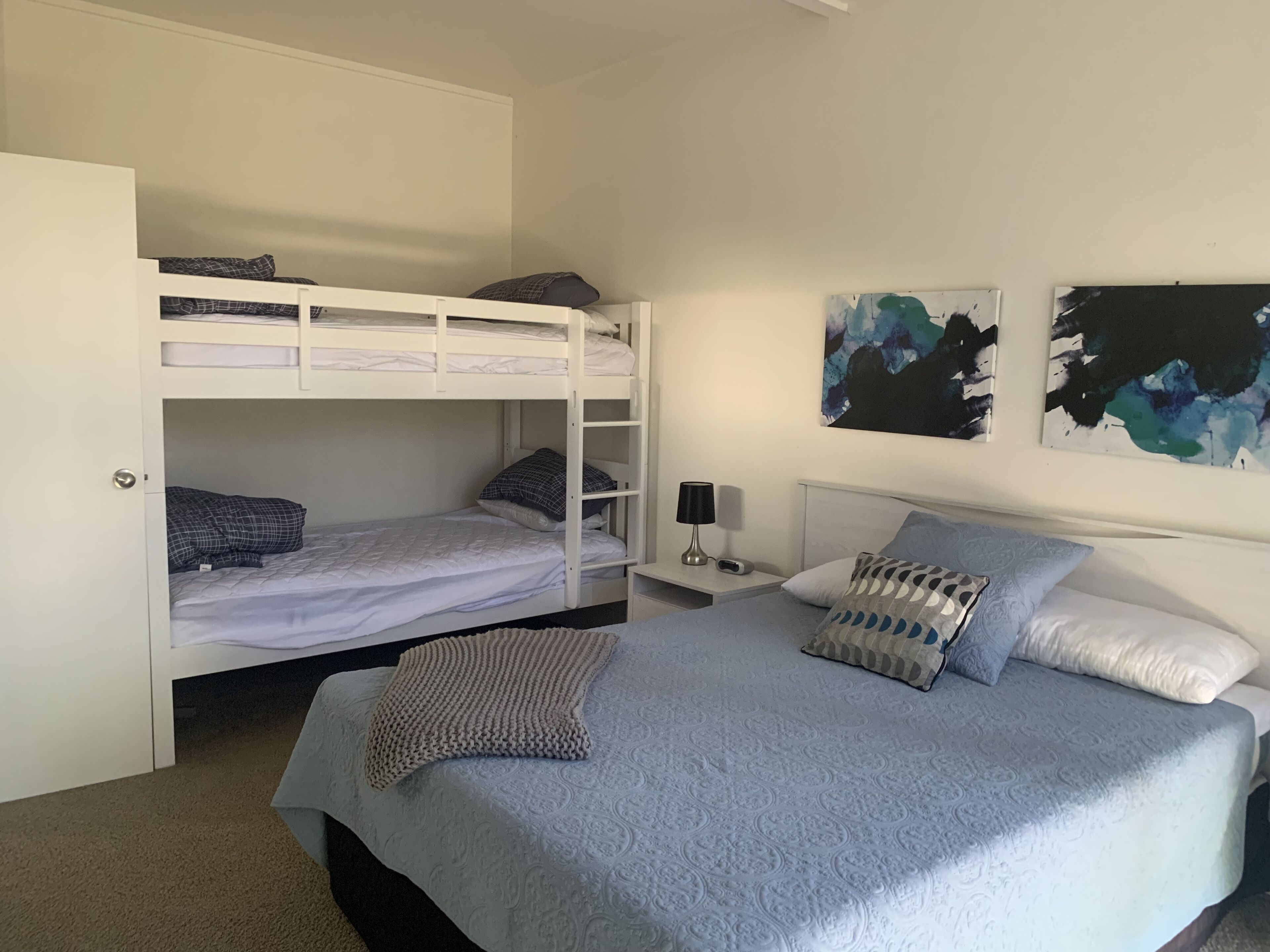A bedroom with a bunk bed, a single bed, a bedside table with a lamp, and abstract wall art.