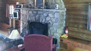 A rustic cabin interior with a stone fireplace, wooden walls, a wooden ceiling, a wooden table, a purple armchair, a television, and a fire extinguisher. - Adirondack Lakeside Log Cabin With Mountain Views (Mountain View)
