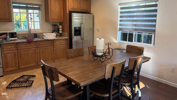 Una cocina con armarios de madera, un refrigerador de acero inoxidable, una mesa de comedor con sillas y un ventanal con persianas.