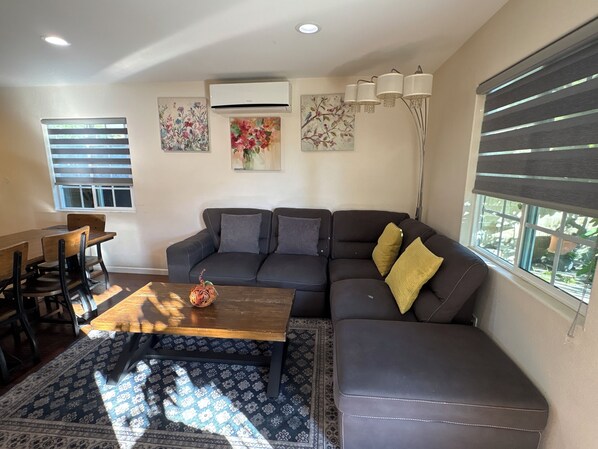 A living room with a sectional sofa, a wooden coffee table, a patterned rug, and a dining area with a table and chairs.