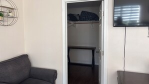 Una habitación con un sofá gris, una puerta blanca que da a un armario con estantes, y un televisor montado en la pared.