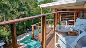 Una terraza de madera con piscina, mobiliario exterior y abundante vegetación.