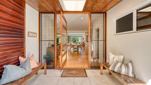 Un interior moderno con un banco de madera, una puerta de cristal de entrada y un área de comedor visible a través de la puerta.