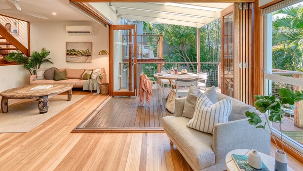 Amplia sala de estar con piso de madera, un sofá, una mesa de centro y una mesa de comedor con sillas. Hay una puerta de vidrio que da a un espacio exterior con vista a árboles.