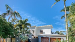 Una casa moderna de dos pisos con techo plano, rodeada de palmeras y un cielo azul despejado.