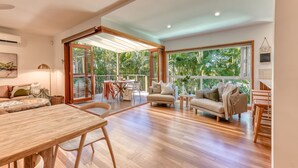Amplia sala de estar con piso de madera, una mesa de comedor, dos sofás y ventanas grandes que ofrecen vistas a exuberante vegetación.