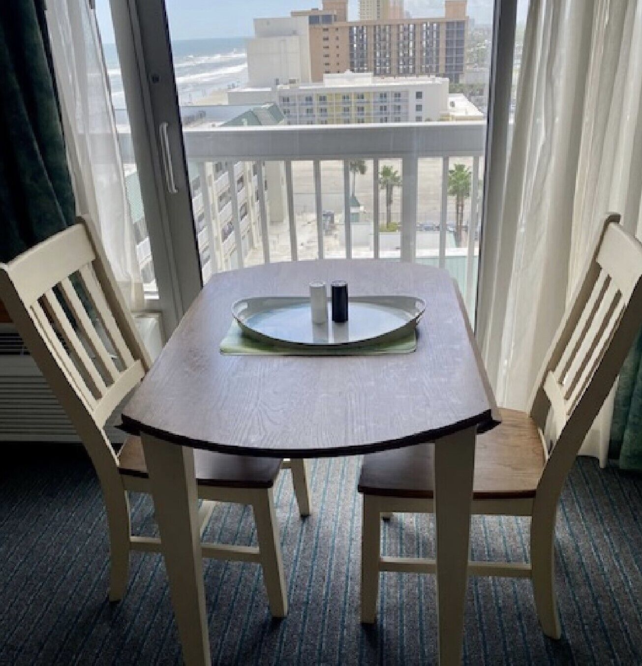 A room with a table and two chairs, a window with a view of buildings and a beach, and a curtain.