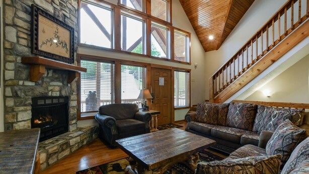 A spacious living room with a stone fireplace, large windows, a wooden ceiling, and a staircase.