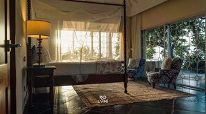 A bedroom with a four-poster bed, a bedside table with a lamp, a patterned rug, and a chair by the window.
