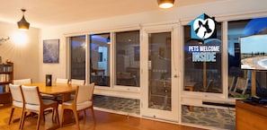 Interior que admet mascotes amb zona de menjador, portes corredisses a un patí i un televisor que mostra una platja.