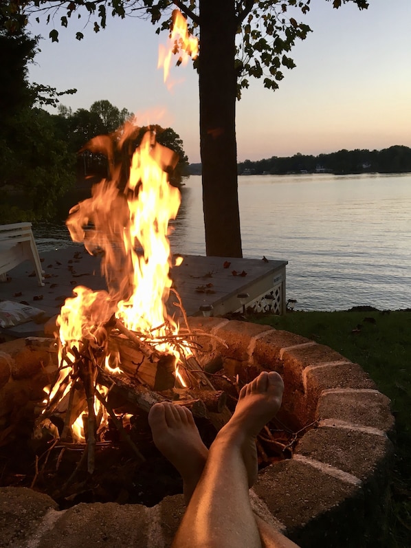 A fire pit with flames, surrounded by a brick fire pit, and a person relaxing with their feet up.
